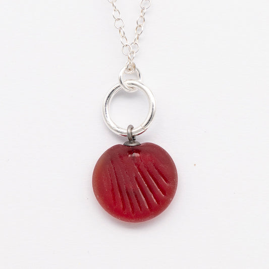 Close up of red frosted glass charm with shell imprint on a plain white background. The charm is frosted, and spiral is shiny. The charm hangs on a large silver jump ring. The necklace is completed with a small silver jump ring and silver chain.