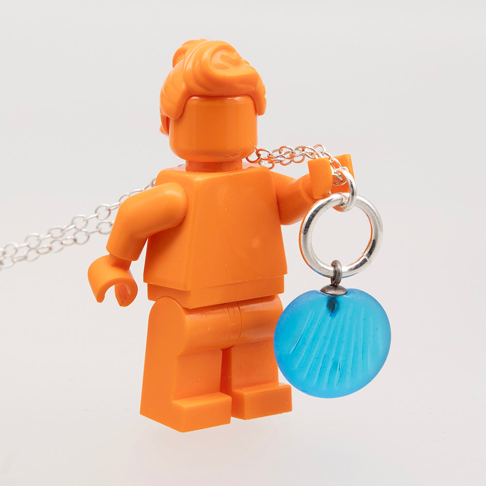 Shell imprint necklace held by orange lego mini figure to show scale. The glass charm is approximately the same height as the legs.