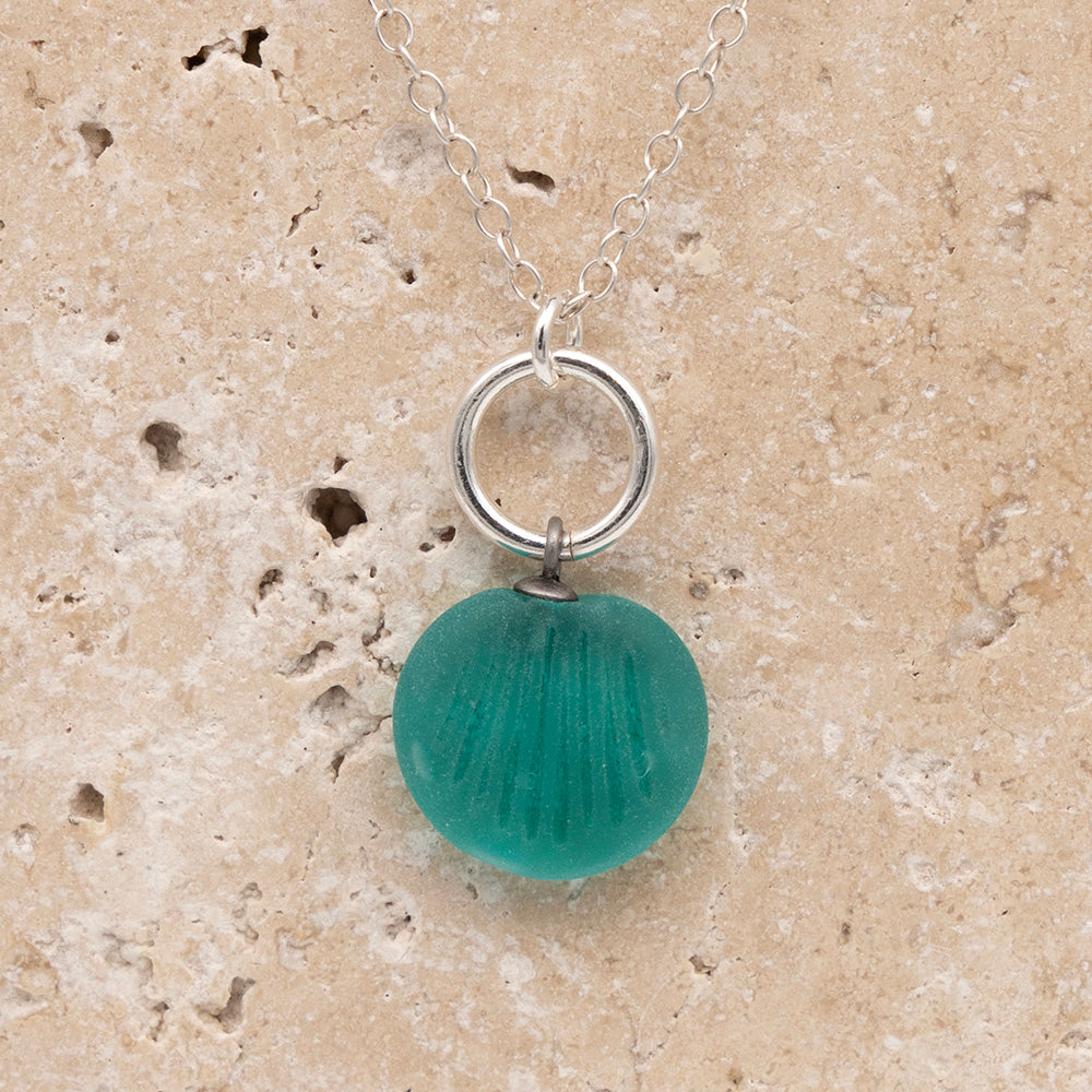 Close up of the front of a teal frosted glass charm with shell imprint on a sandstone background. The charm is frosted, and spiral is shiny. The charm hangs on a large silver jump ring. The necklace is completed with a small silver jump ring and silver chain.