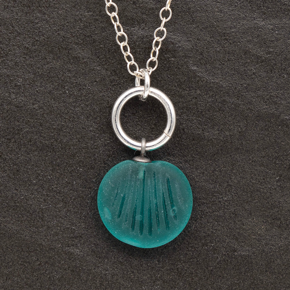 Close up of a teal frosted glass charm with shell imprint on a dark slate background. The charm is frosted, and spiral is shiny. The charm hangs on a large silver jump ring. The necklace is completed with a small silver jump ring and silver chain.