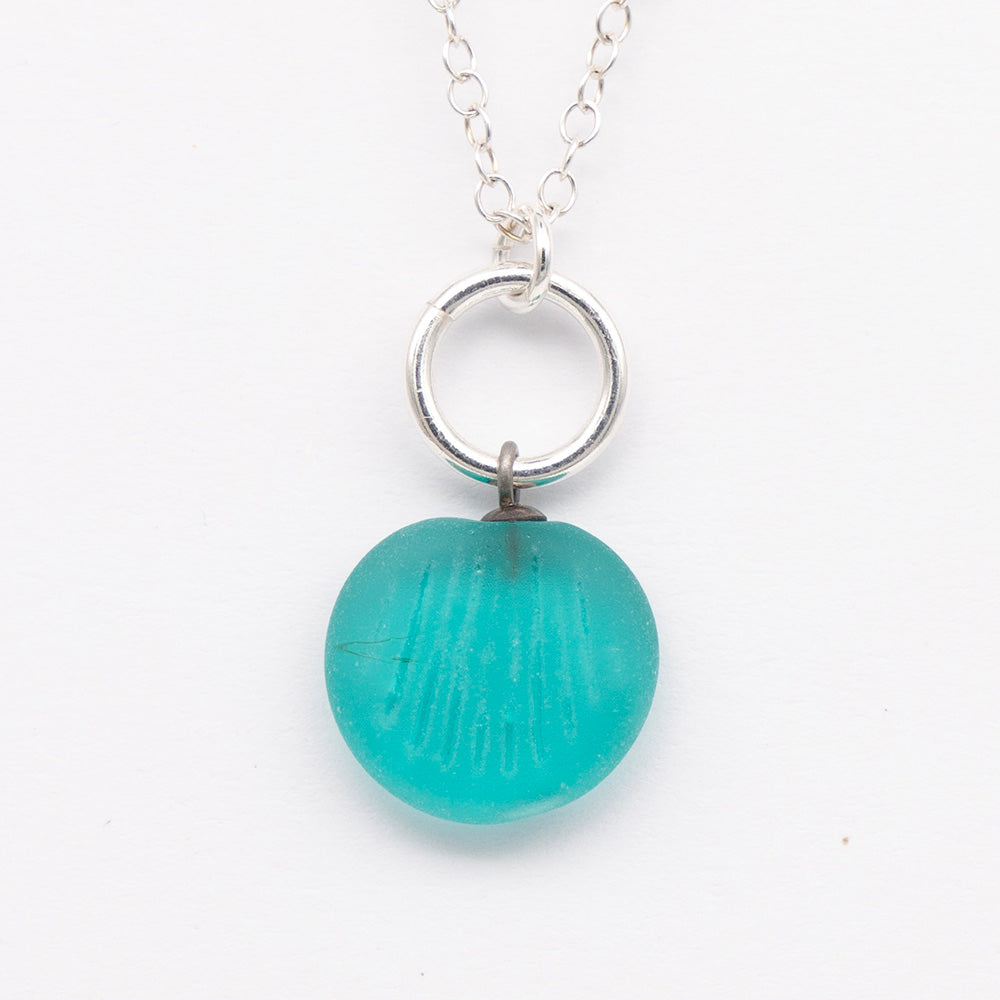 Close up of teal frosted glass charm with shell imprint on a plain white background. The charm is frosted, and spiral is shiny. The charm hangs on a large silver jump ring. The necklace is completed with a small silver jump ring and silver chain.