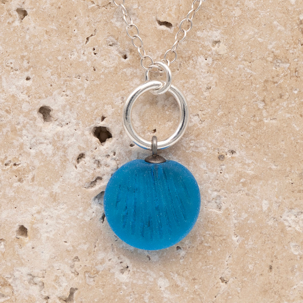 Close up of the front of a turquoise frosted glass charm with shell imprint on a sandstone background. The charm is frosted, and spiral is shiny. The charm hangs on a large silver jump ring. The necklace is completed with a small silver jump ring and silver chain.