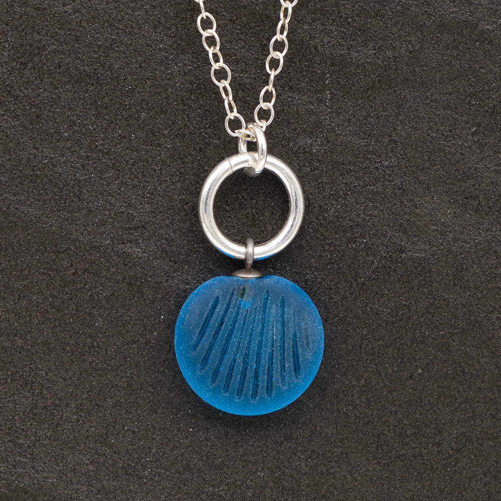 Close up of a turquoise frosted glass charm with shell imprint on a dark slate background. The charm is frosted, and spiral is shiny. The charm hangs on a large silver jump ring. The necklace is completed with a small silver jump ring and silver chain.