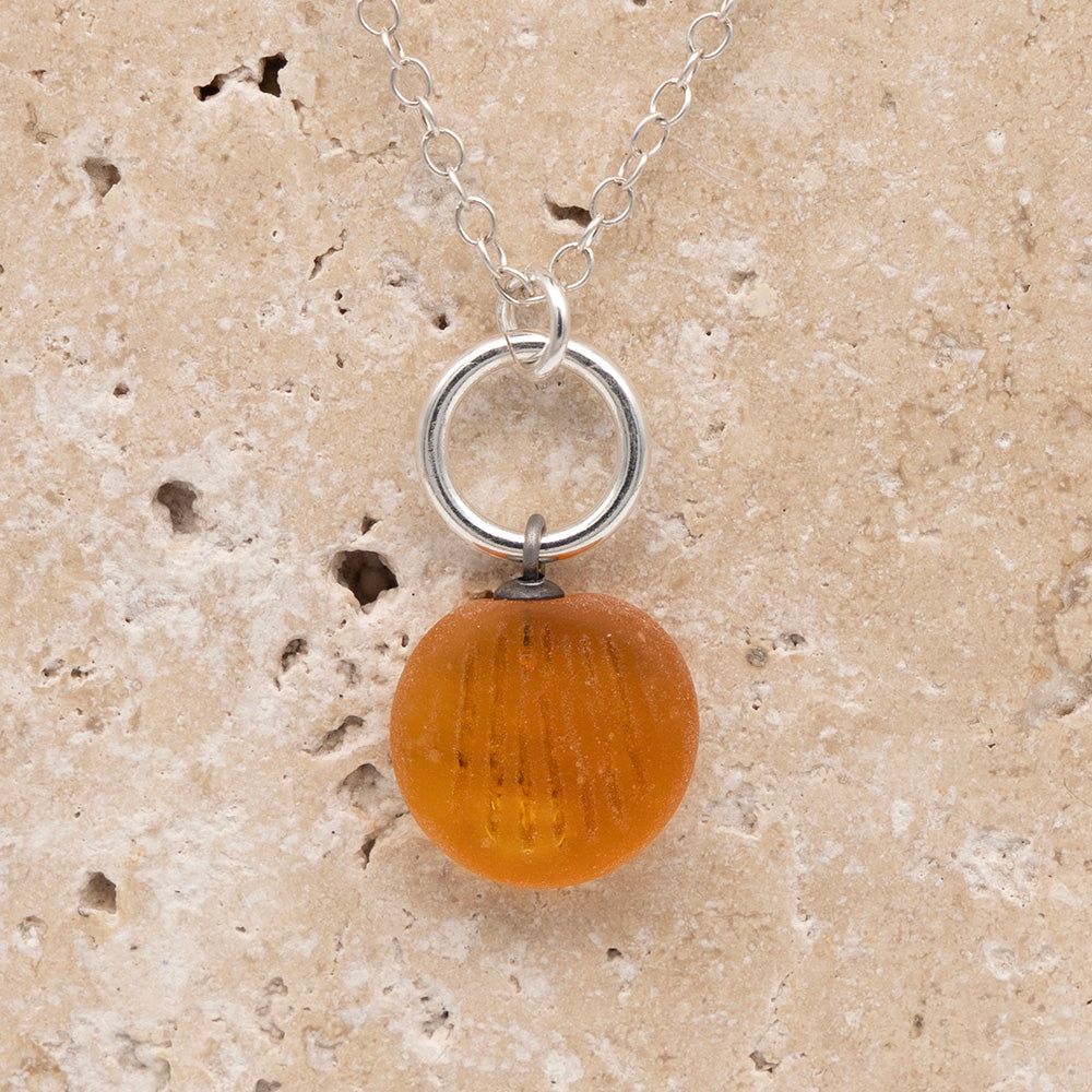 Close up of the front of a yellow frosted glass charm with shell imprint on a sandstone background. The charm is frosted, and spiral is shiny. The charm hangs on a large silver jump ring. The necklace is completed with a small silver jump ring and silver chain.