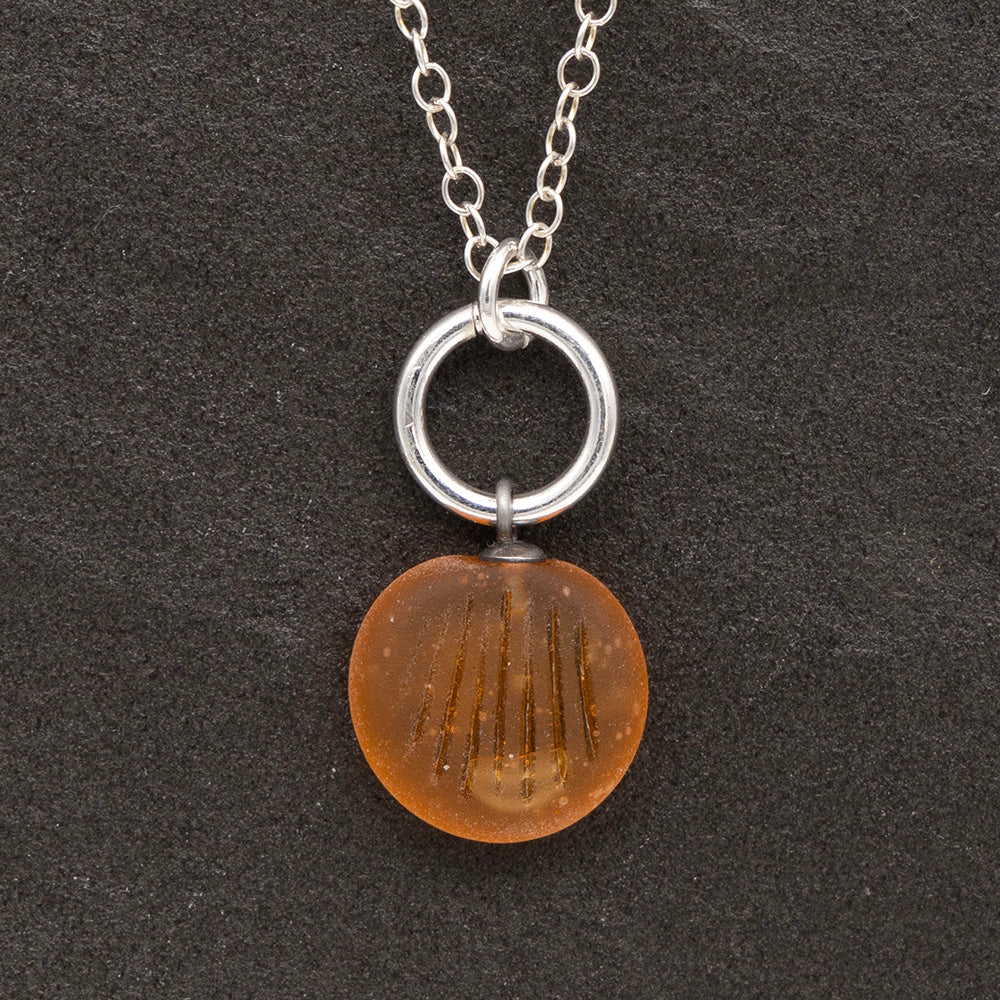 Close up of a yellow frosted glass charm with shell imprint on a dark slate background. The charm is frosted, and spiral is shiny. The charm hangs on a large silver jump ring. The necklace is completed with a small silver jump ring and silver chain.