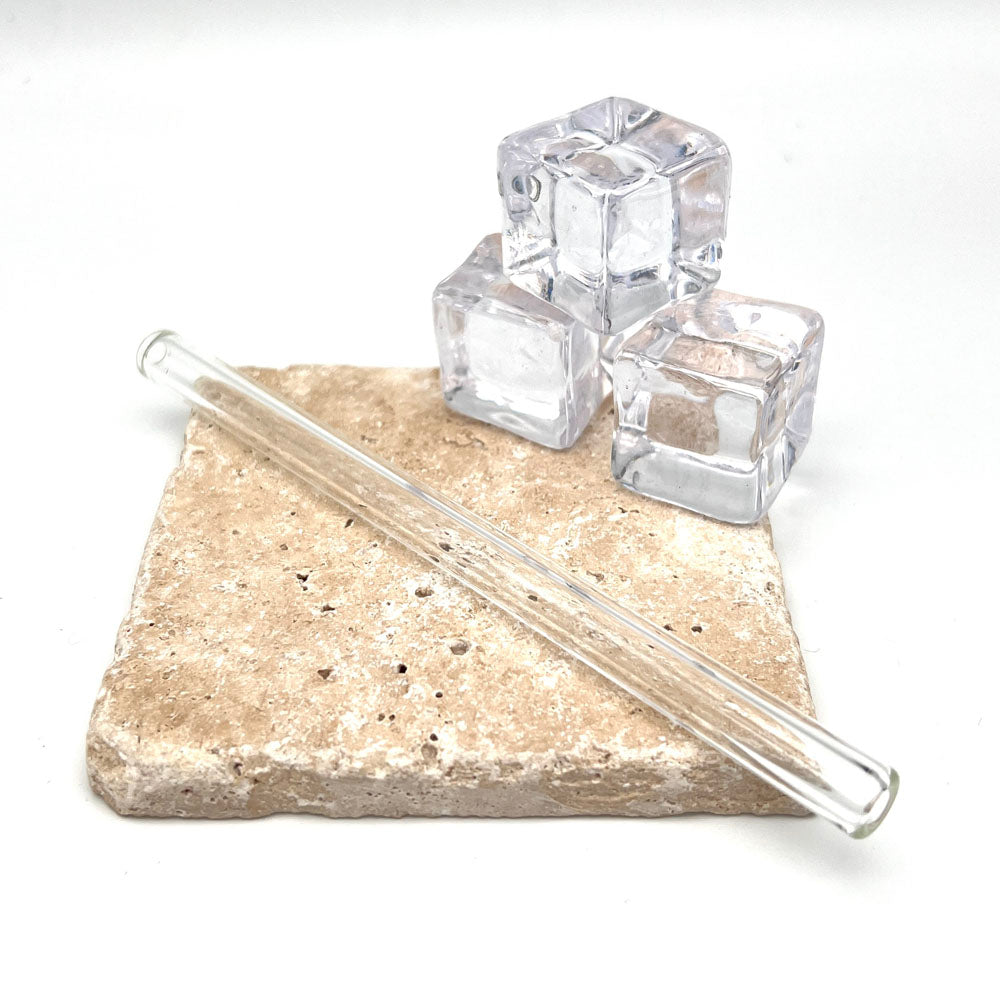 A short clear glass straw sits on a sandstone tile in front of three ice cubes