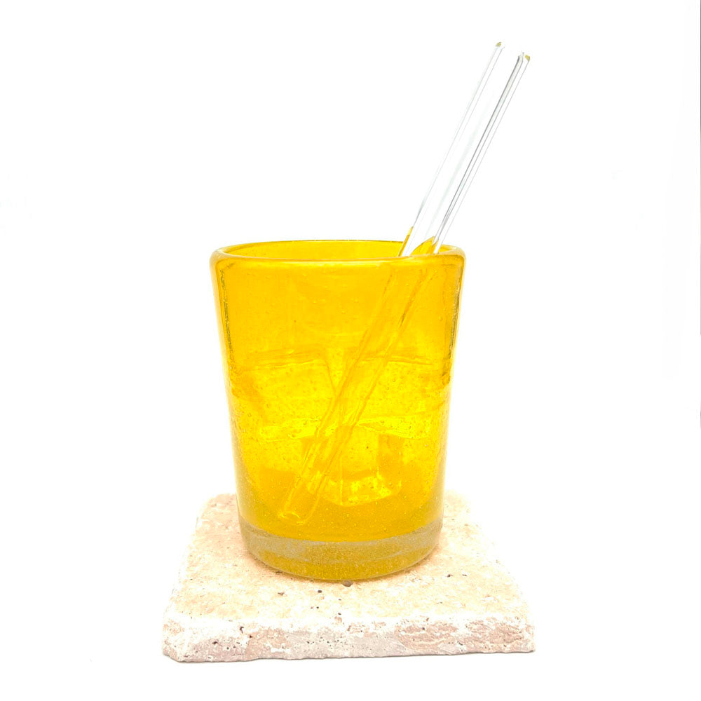 A short clar glass straw sitting in a yellow tumbler filled with ice. The tumbler sits on a sandstone tile.