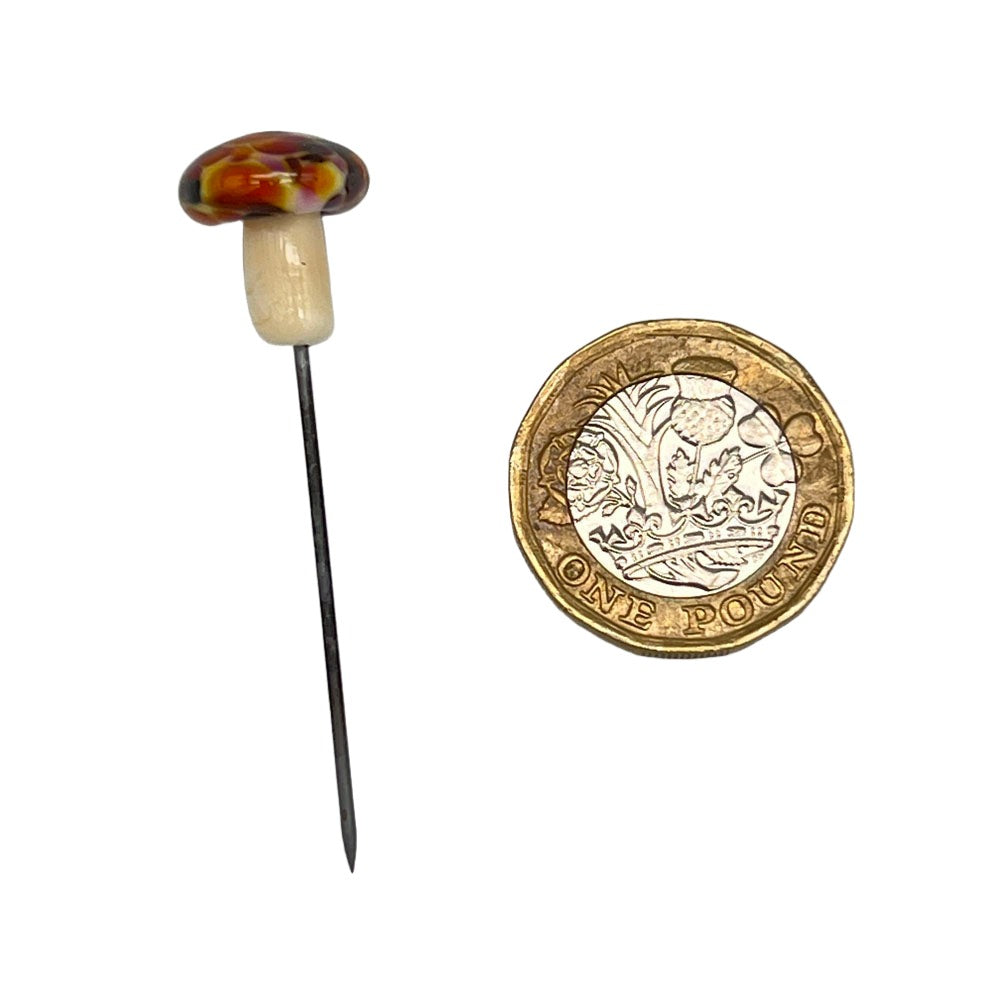 Speckled mushroom pin next to a pound coin: A handmade glass mushroom pin with a speckled cap featuring shades of berry, brown and orange, displayed next to a one-pound coin for size comparison.