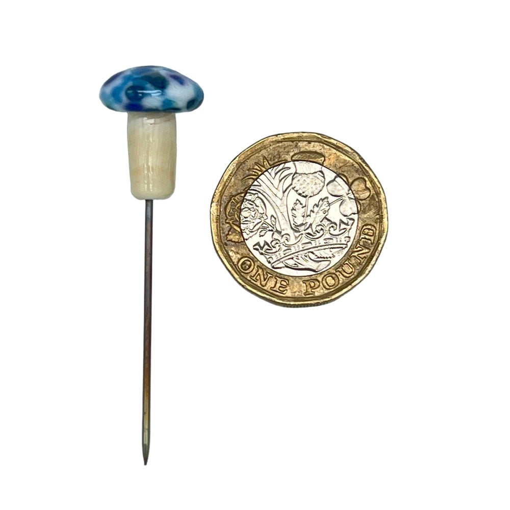 Speckled mushroom pin with blue cap next to a pound coin: A handmade glass mushroom pin with a blue and white speckled cap, shown alongside a one-pound coin for size reference.