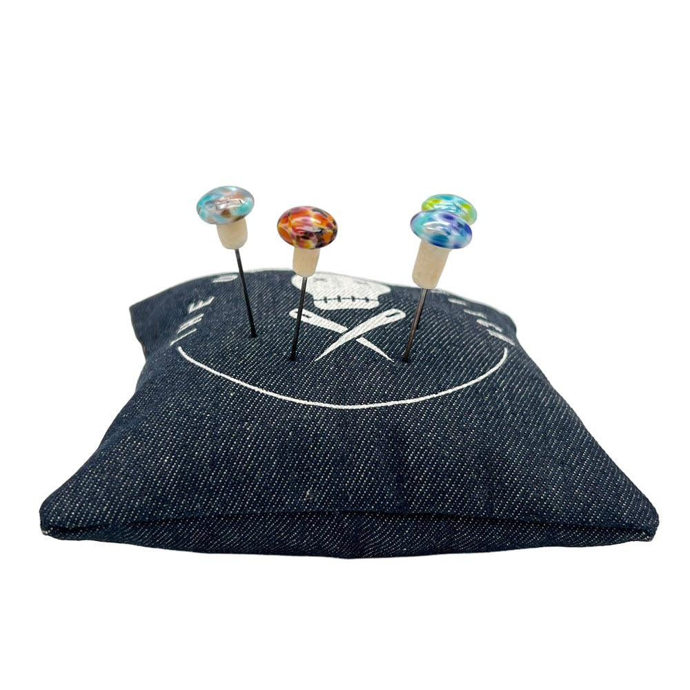 Speckled mushroom pins on a pin cushion: Four colorful handmade glass mushroom pins inserted into a dark blue pin cushion with a white skull and crossbones design.