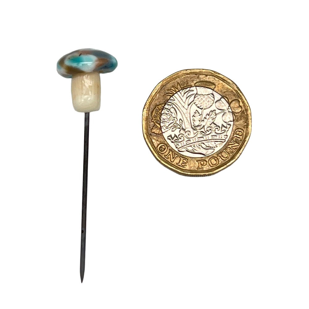 Speckled mushroom pin with turquoise cap next to a pound coin: A handmade glass mushroom pin with a turquoise and brown speckled cap, placed beside a one-pound coin for size comparison.