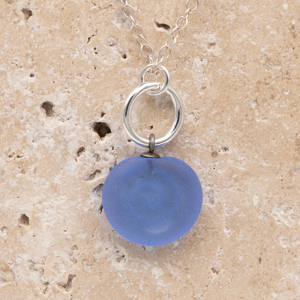 Close up of the back of a blue frosted glass charm with swirl imprint on a sandstone background. The whole charm is frosted, and spiral is plain frosted glass. The charm hangs on a large silver jump ring. The necklace is completed with a small silver jump ring and silver chain.