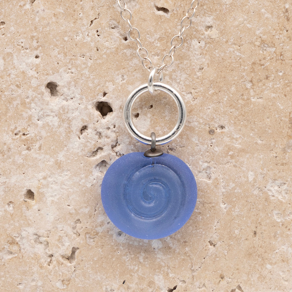 Close up of the front  of a blue frosted glass charm with swirl imprint on a sandstone background. The charm is frosted and spiral is shiny. The charm hangs on a large silver jump ring. The necklace is completed with a small silver jump ring and silver chain.