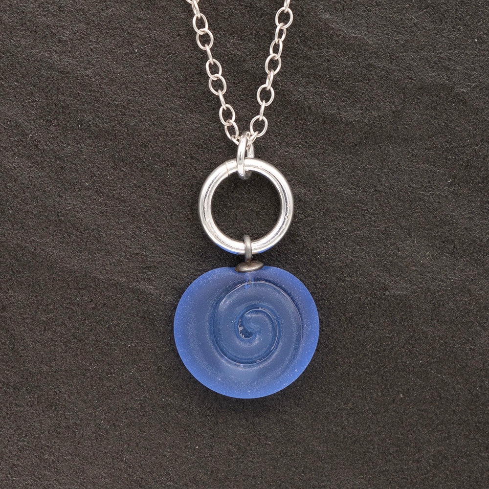 Close up of blue frosted glass charm with swirl imprint on a dark slate background. The charm is frosted and spiral is shiny. The charm hangs on a large silver jump ring. The necklace is completed with a small silver jump ring and silver chain.