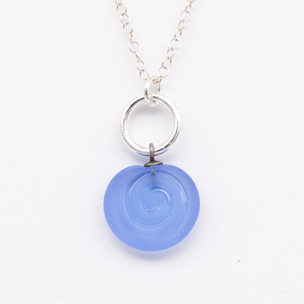 Close up of blue frosted glass charm with swirl imprint. The charm is frosted and spiral is shiny. The charm hangs on a large silver jump ring. The necklace is completed with a small silver jump ring and silver chain.