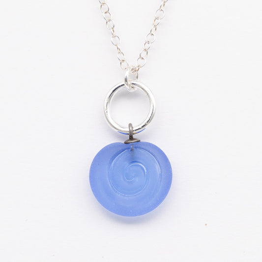 Close up of blue frosted glass charm with swirl imprint. The charm is frosted and spiral is shiny. The charm hangs on a large silver jump ring. The necklace is completed with a small silver jump ring and silver chain.