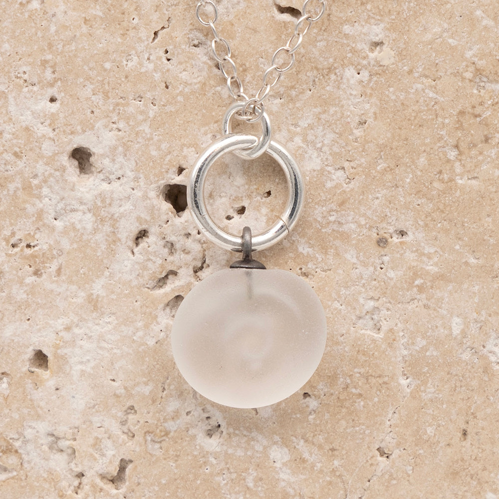 Close up of the back of a clear frosted glass charm with swirl imprint on a sandstone background. The whole charm is frosted, and spiral is plain frosted glass. The charm hangs on a large silver jump ring. The necklace is completed with a small silver jump ring and silver chain.