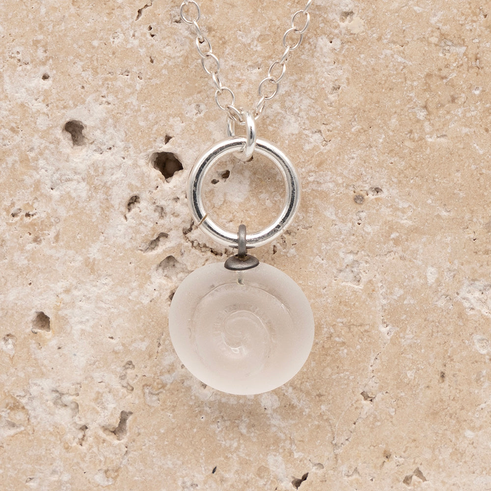 Close up of the front  of a clear frosted glass charm with swirl imprint on a sandstone background. The charm is frosted and spiral is shiny. The charm hangs on a large silver jump ring. The necklace is completed with a small silver jump ring and silver chain.