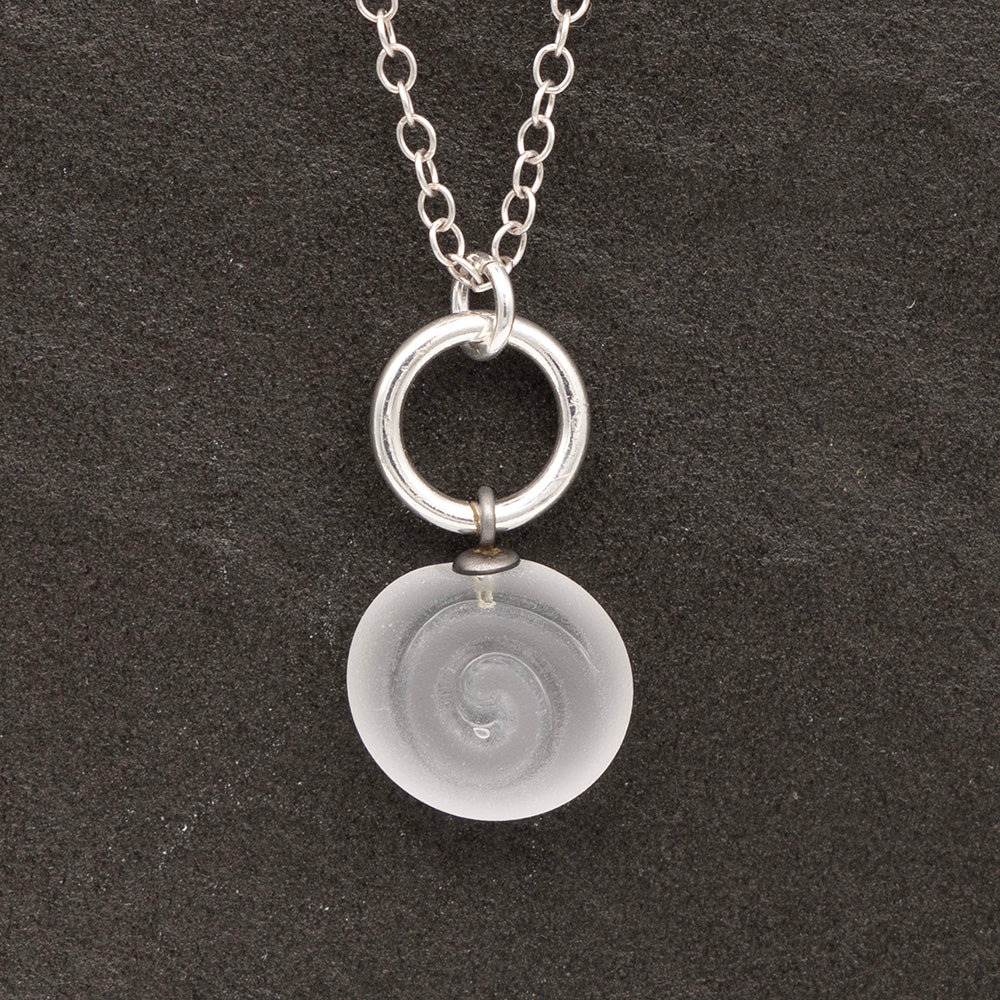 Close up of clear frosted glass charm with swirl imprint on a dark slate background. The charm is frosted and spiral is shiny. The charm hangs on a large silver jump ring. The necklace is completed with a small silver jump ring and silver chain.