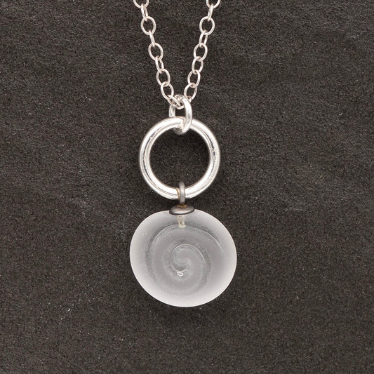 Close up of clear frosted glass charm with swirl imprint on a dark slate background. The charm is frosted and spiral is shiny. The charm hangs on a large silver jump ring. The necklace is completed with a small silver jump ring and silver chain.