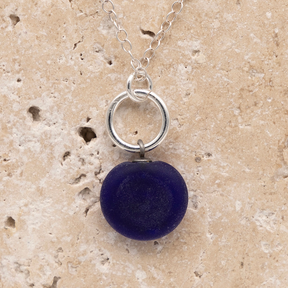 Close up of the back of a dark blue frosted glass charm with swirl imprint on a sandstone background. The whole charm is frosted, and spiral is plain frosted glass. The charm hangs on a large silver jump ring. The necklace is completed with a small silver jump ring and silver chain.