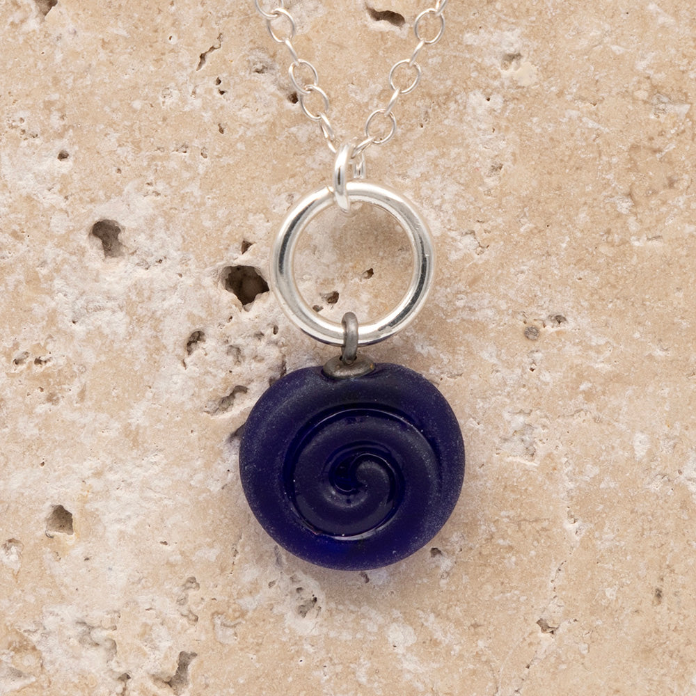 Close up of the front  of a dark blue frosted glass charm with swirl imprint on a sandstone background. The charm is frosted and spiral is shiny. The charm hangs on a large silver jump ring. The necklace is completed with a small silver jump ring and silver chain.