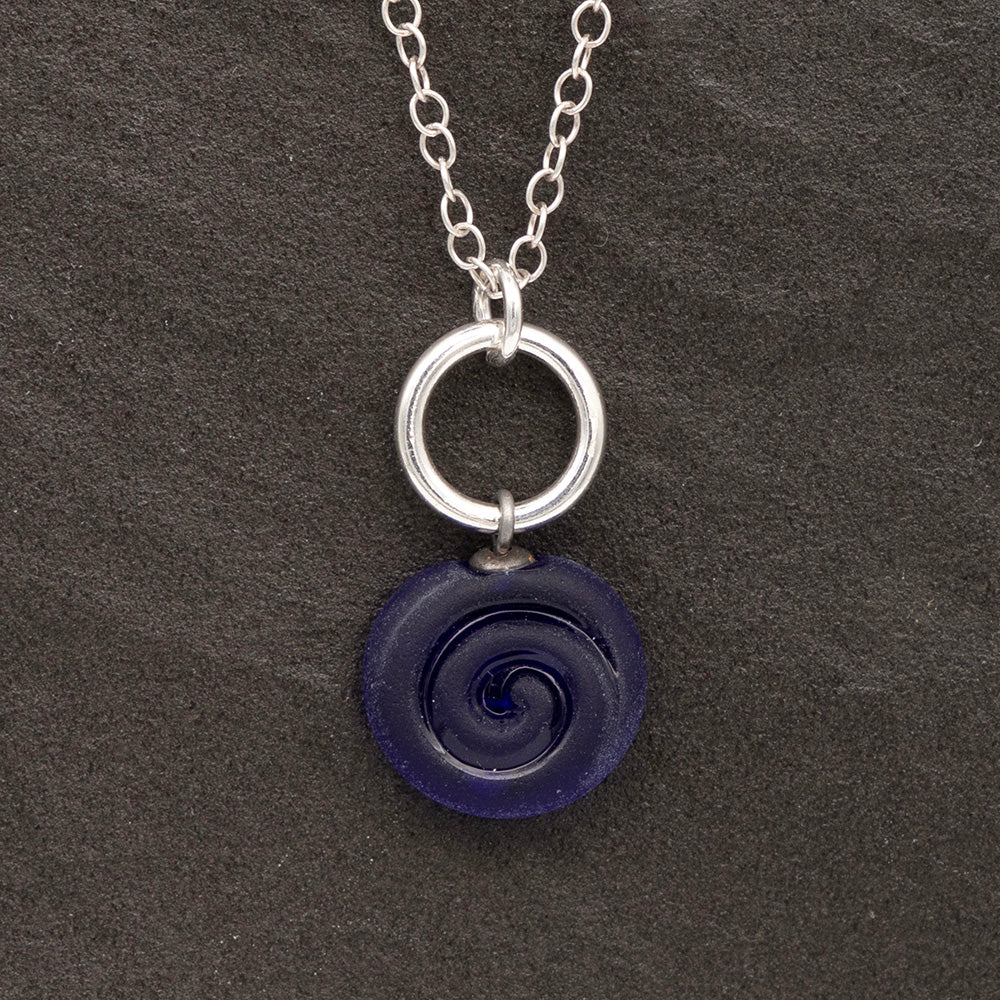 Close up of dark blue frosted glass charm with swirl imprint on a dark slate background. The charm is frosted and spiral is shiny. The charm hangs on a large silver jump ring. The necklace is completed with a small silver jump ring and silver chain.