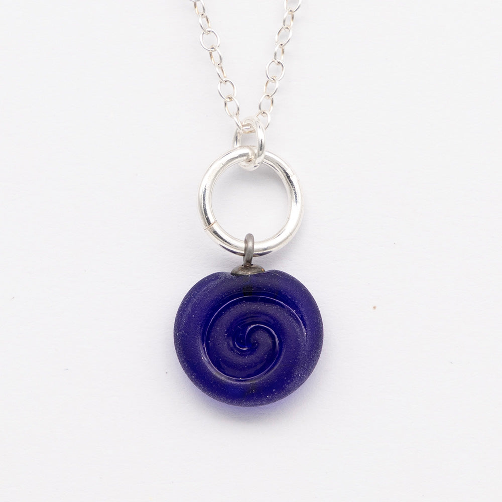 Close up of dark blue frosted glass charm with swirl imprint. The charm is frosted and spiral is shiny. The charm hangs on a large silver jump ring. The necklace is completed with a small silver jump ring and silver chain.