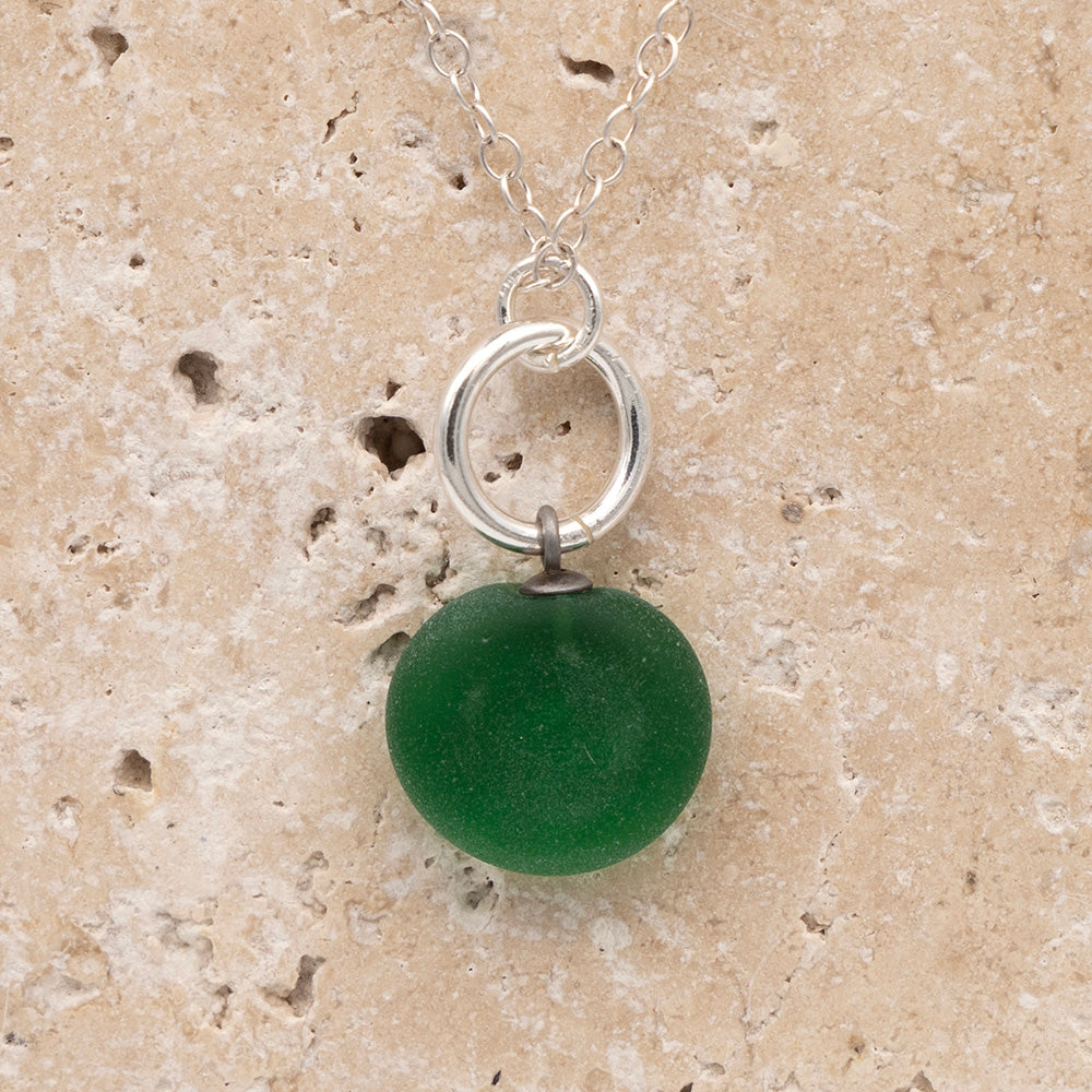 Close up of the back of a green frosted glass charm with swirl imprint on a sandstone background. The whole charm is frosted, and spiral is plain frosted glass. The charm hangs on a large silver jump ring. The necklace is completed with a small silver jump ring and silver chain.