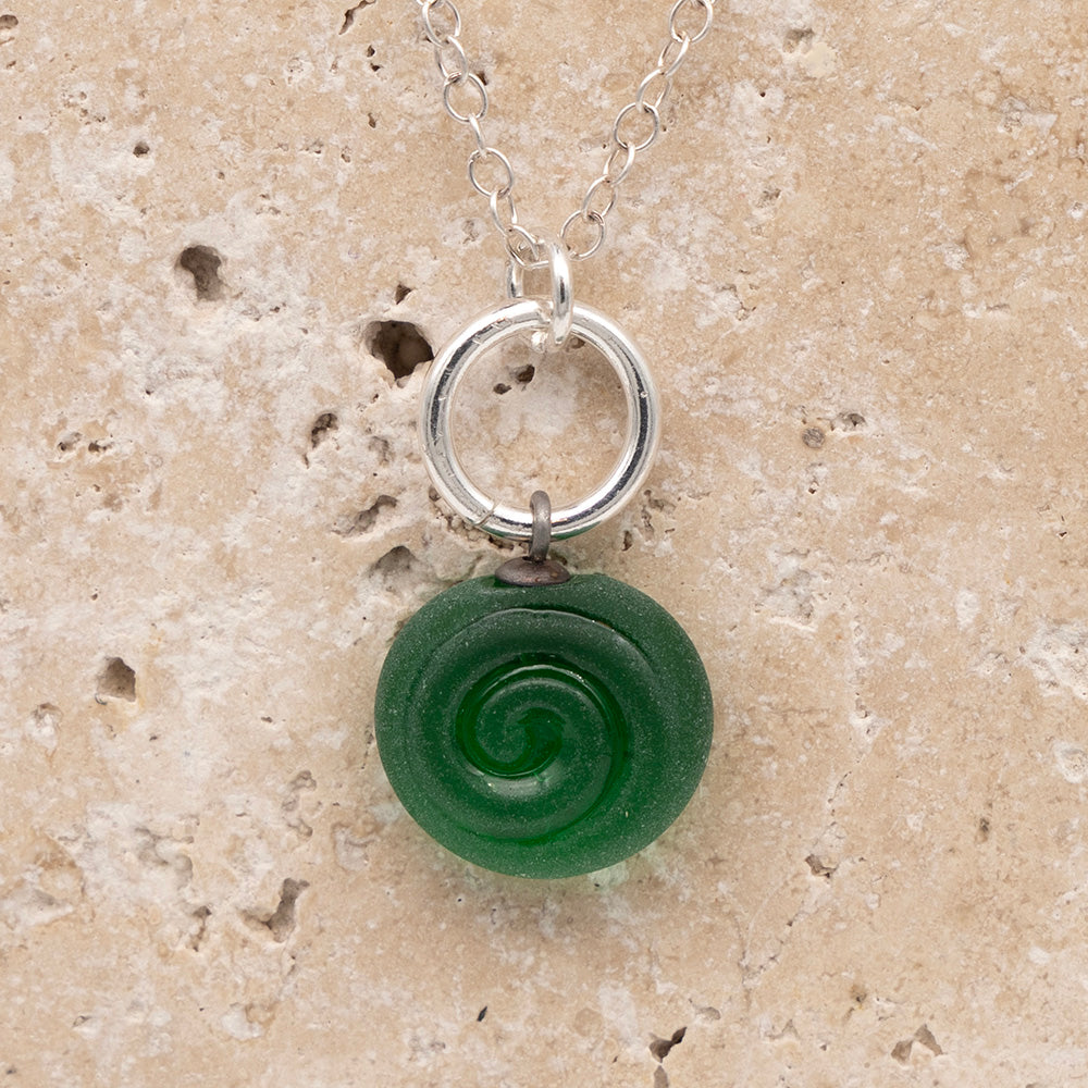 Close up of the front  of a green frosted glass charm with swirl imprint on a sandstone background. The charm is frosted and spiral is shiny. The charm hangs on a large silver jump ring. The necklace is completed with a small silver jump ring and silver chain.