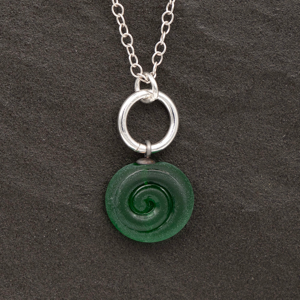 Close up of green frosted glass charm with swirl imprint on a dark slate background. The charm is frosted and spiral is shiny. The charm hangs on a large silver jump ring. The necklace is completed with a small silver jump ring and silver chain.