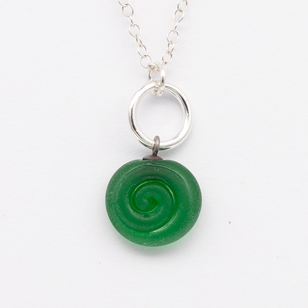 Close up of green frosted glass charm with swirl imprint. The charm is frosted and spiral is shiny. The charm hangs on a large silver jump ring. The necklace is completed with a small silver jump ring and silver chain.