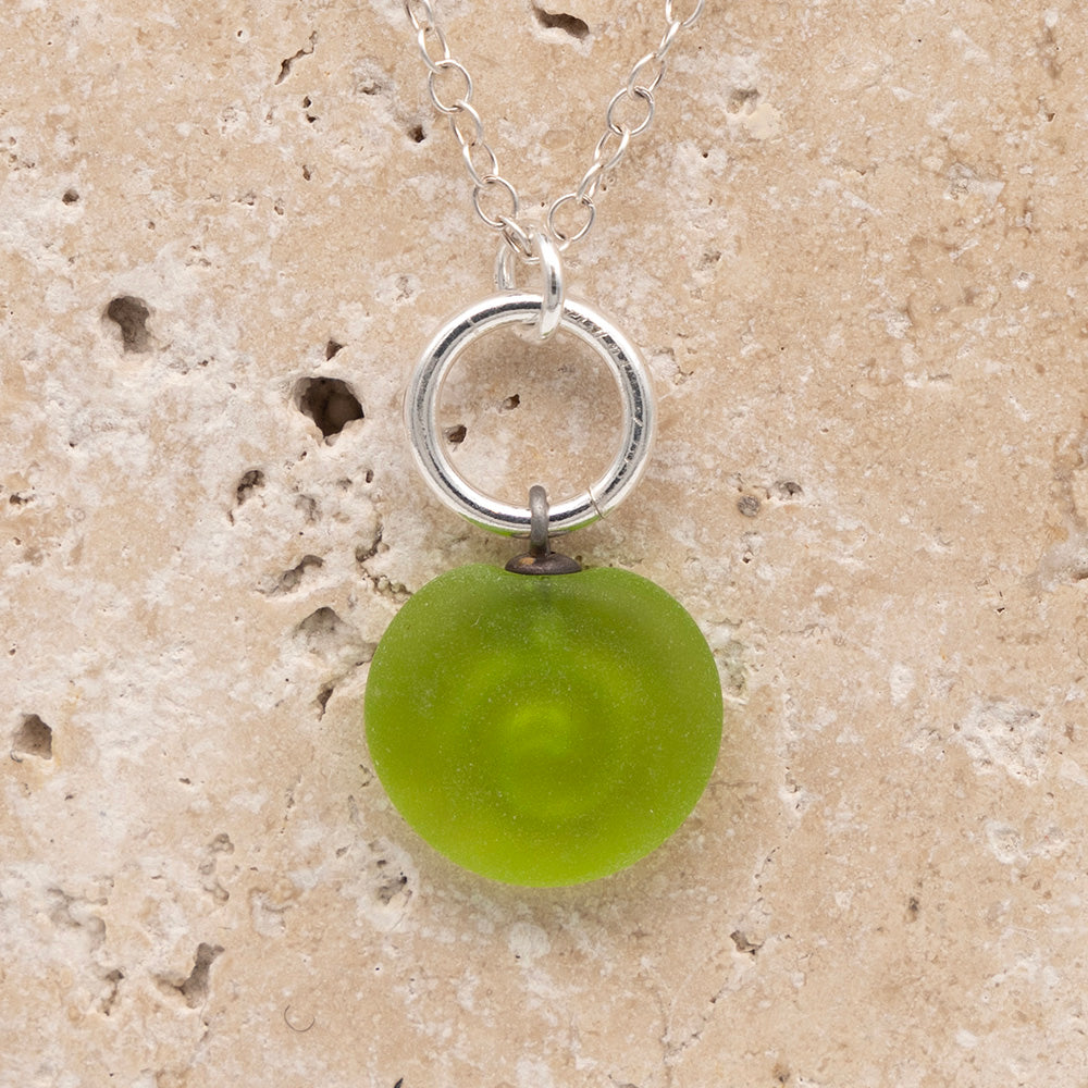 Close up of the back of a lime frosted glass charm with swirl imprint on a sandstone background. The whole charm is frosted, and spiral is plain frosted glass. The charm hangs on a large silver jump ring. The necklace is completed with a small silver jump ring and silver chain.