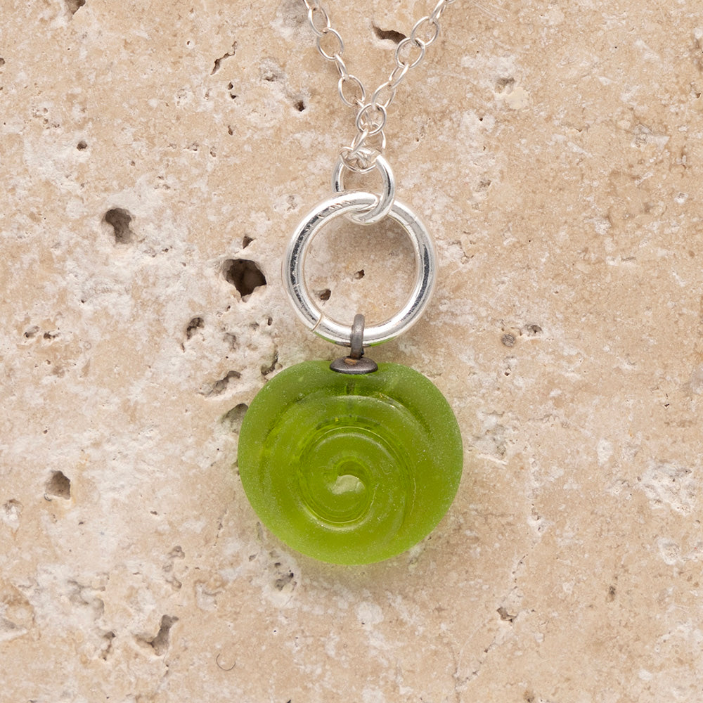 Close up of the front  of a lime frosted glass charm with swirl imprint on a sandstone background. The charm is frosted and spiral is shiny. The charm hangs on a large silver jump ring. The necklace is completed with a small silver jump ring and silver chain.