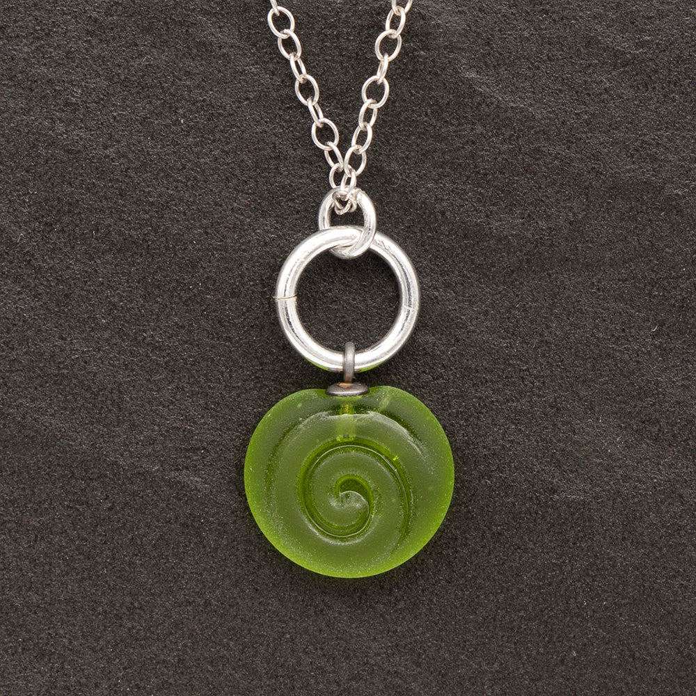 Close up of lime frosted glass charm with swirl imprint on a dark slate background. The charm is frosted and spiral is shiny. The charm hangs on a large silver jump ring. The necklace is completed with a small silver jump ring and silver chain.