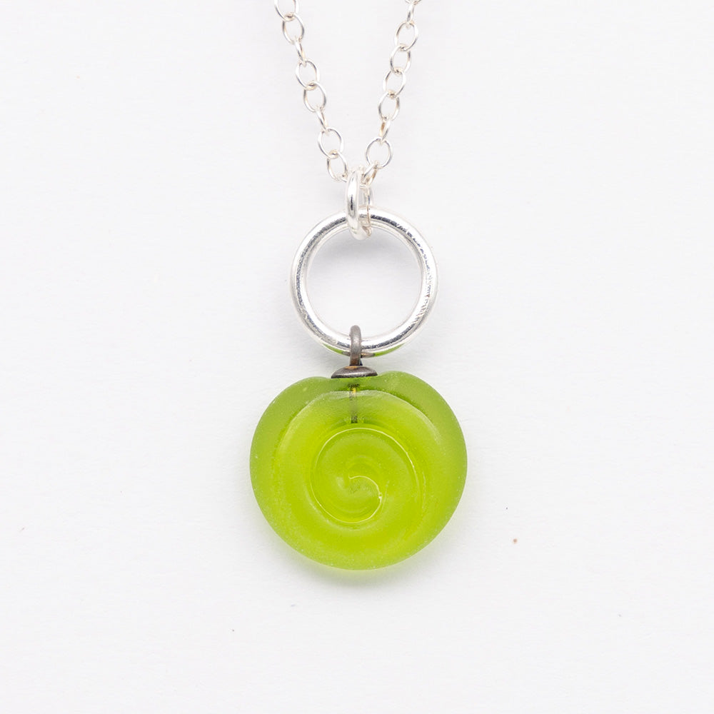 Close up of lime frosted glass charm with swirl imprint. The charm is frosted and spiral is shiny. The charm hangs on a large silver jump ring. The necklace is completed with a small silver jump ring and silver chain.