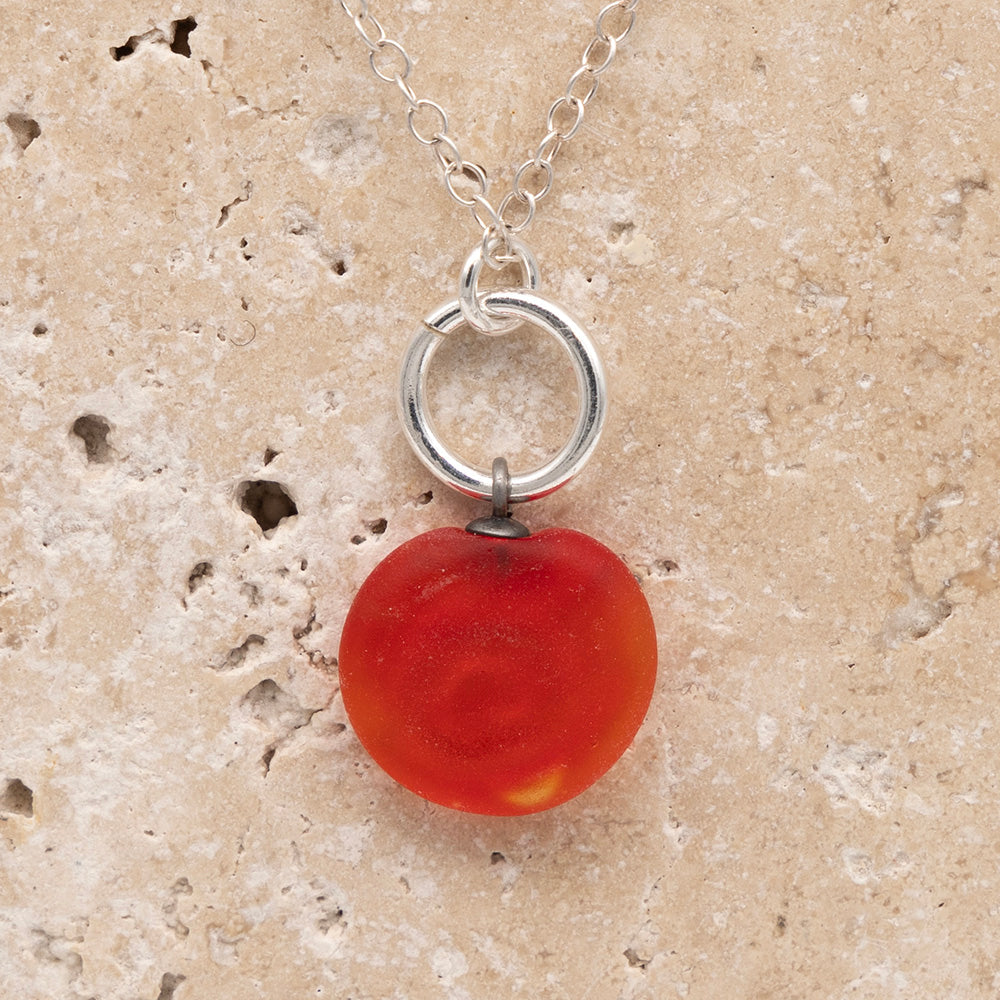 Close up of the back of an orange frosted glass charm with swirl imprint on a sandstone background. The whole charm is frosted, and spiral is plain frosted glass. The charm hangs on a large silver jump ring. The necklace is completed with a small silver jump ring and silver chain.