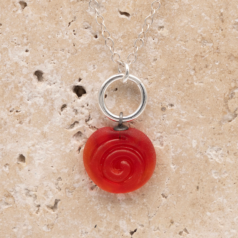 Close up of the front  of a orange frosted glass charm with swirl imprint on a sandstone background. The charm is frosted and spiral is shiny. The charm hangs on a large silver jump ring. The necklace is completed with a small silver jump ring and silver chain.