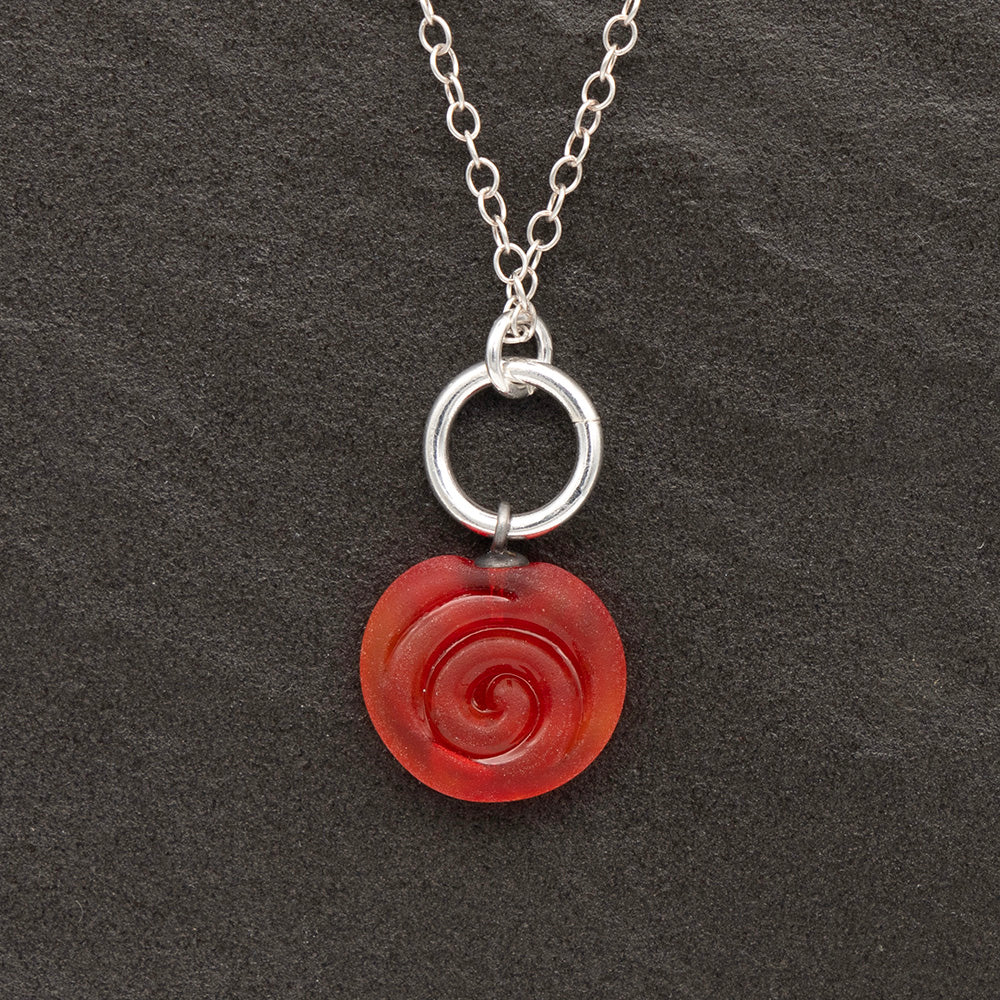 Close up of orange frosted glass charm with swirl imprint on a dark slate background. The charm is frosted and spiral is shiny. The charm hangs on a large silver jump ring. The necklace is completed with a small silver jump ring and silver chain.