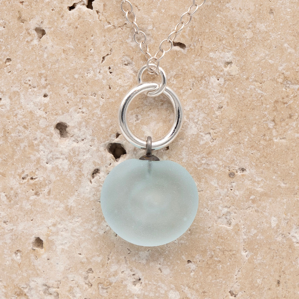 Close up of the back of a pale blue frosted glass charm with swirl imprint on a sandstone background. The whole charm is frosted, and spiral is plain frosted glass. The charm hangs on a large silver jump ring. The necklace is completed with a small silver jump ring and silver chain.