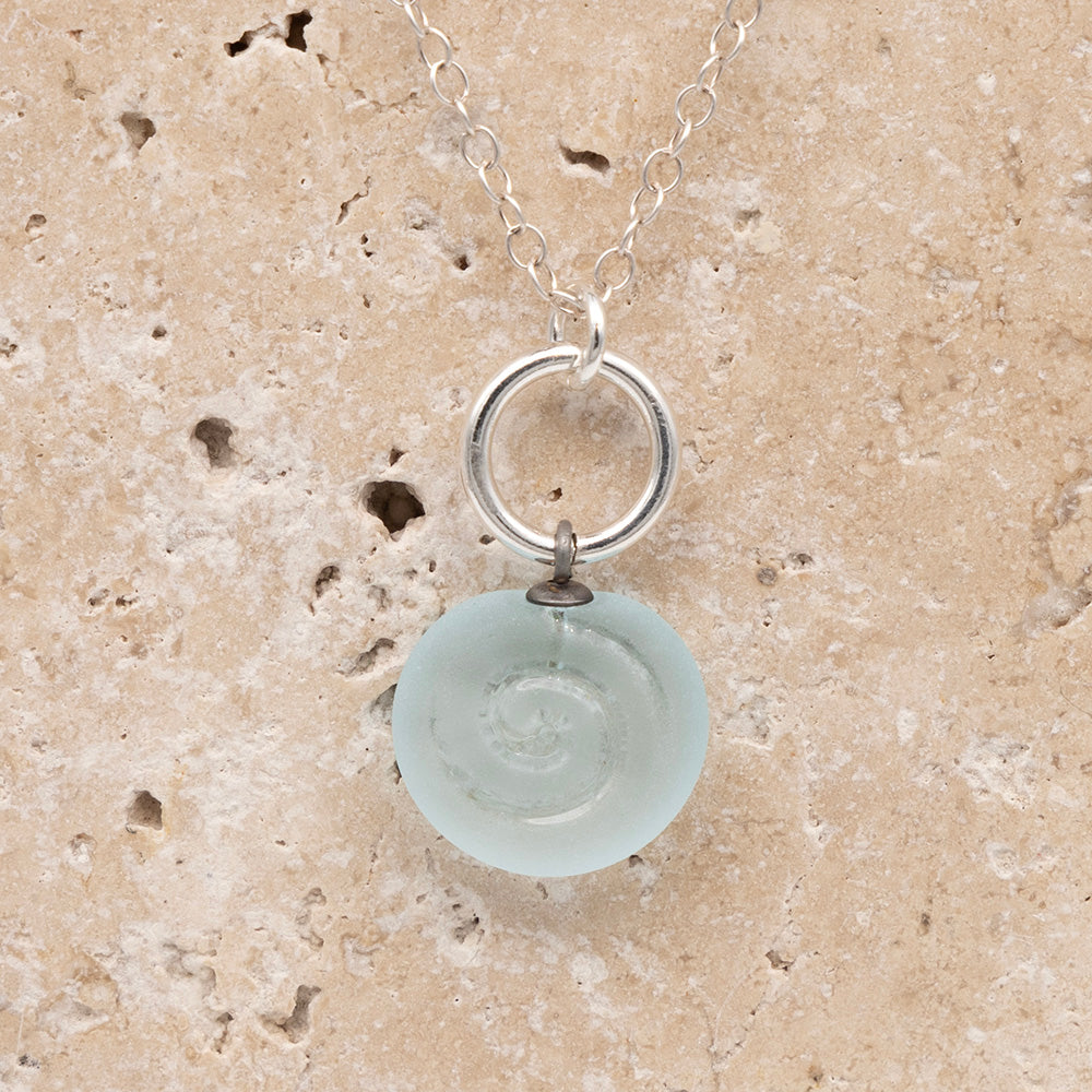 Close up of the front  of a pale blue frosted glass charm with swirl imprint on a sandstone background. The charm is frosted and spiral is shiny. The charm hangs on a large silver jump ring. The necklace is completed with a small silver jump ring and silver chain.