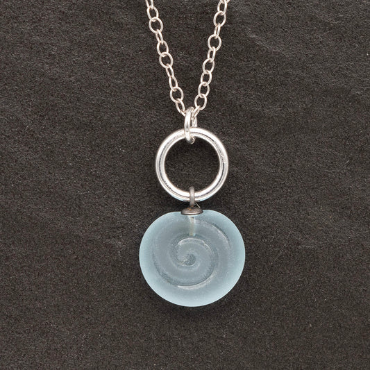 Close up of pale blue frosted glass charm with swirl imprint on a dark slate background. The charm is frosted and spiral is shiny. The charm hangs on a large silver jump ring. The necklace is completed with a small silver jump ring and silver chain.