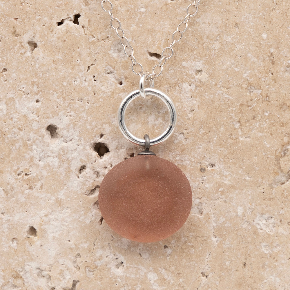 Close up of the back of a pink frosted glass charm with swirl imprint on a sandstone background. The whole charm is frosted, and spiral is plain frosted glass. The charm hangs on a large silver jump ring. The necklace is completed with a small silver jump ring and silver chain.