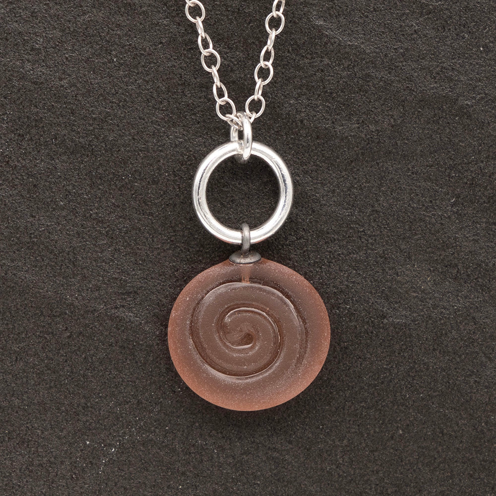 Close up of pink frosted glass charm with swirl imprint on a dark slate background. The charm is frosted and spiral is shiny. The charm hangs on a large silver jump ring. The necklace is completed with a small silver jump ring and silver chain.