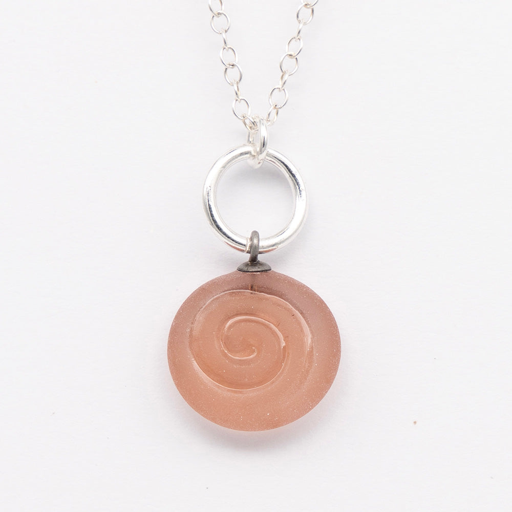 Close up of pink frosted glass charm with swirl imprint. The charm is frosted and spiral is shiny. The charm hangs on a large silver jump ring. The necklace is completed with a small silver jump ring and silver chain.