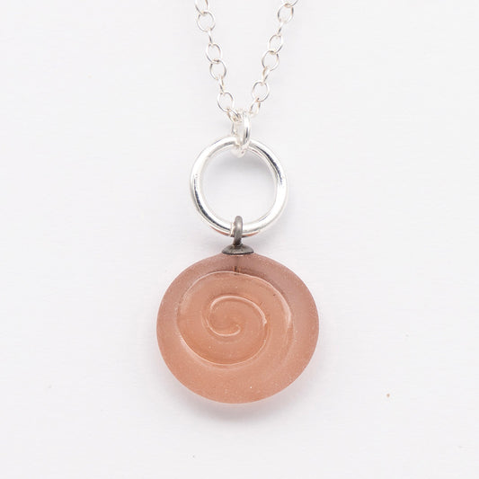 Close up of pink frosted glass charm with swirl imprint. The charm is frosted and spiral is shiny. The charm hangs on a large silver jump ring. The necklace is completed with a small silver jump ring and silver chain.