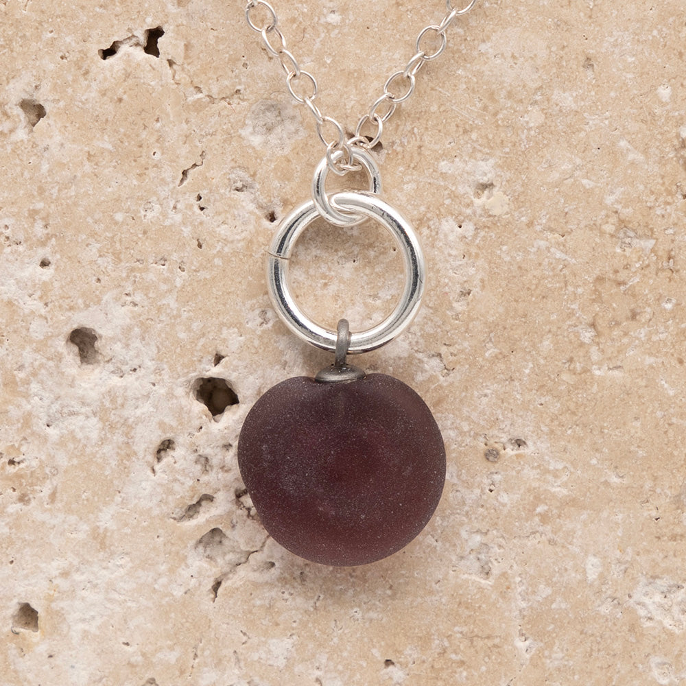 Close up of the back of a purple frosted glass charm with swirl imprint on a sandstone background. The whole charm is frosted, and spiral is plain frosted glass. The charm hangs on a large silver jump ring. The necklace is completed with a small silver jump ring and silver chain.