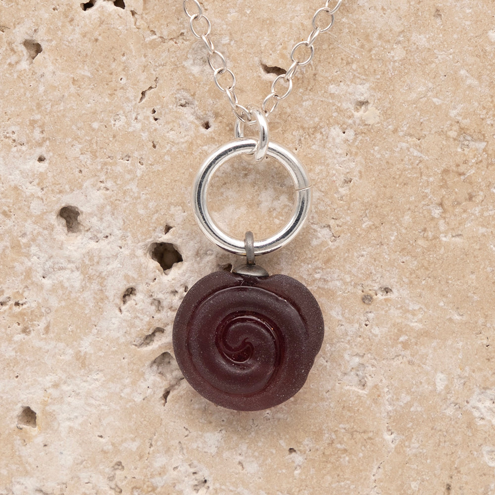 Close up of the front  of a purple frosted glass charm with swirl imprint on a sandstone background. The charm is frosted and spiral is shiny. The charm hangs on a large silver jump ring. The necklace is completed with a small silver jump ring and silver chain.