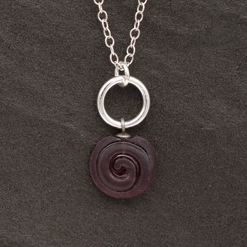 Close up of purple frosted glass charm with swirl imprint on a dark slate background. The charm is frosted and spiral is shiny. The charm hangs on a large silver jump ring. The necklace is completed with a small silver jump ring and silver chain.