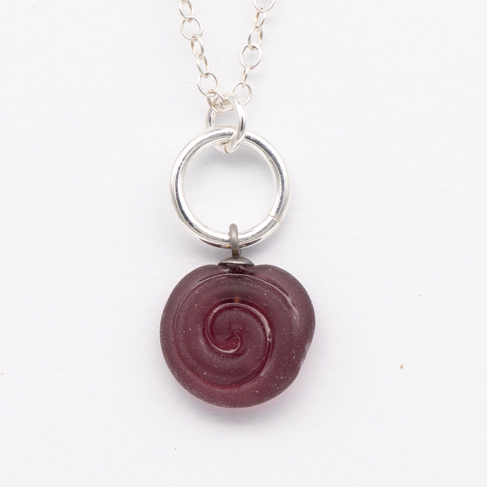 Close up of purple frosted glass charm with swirl imprint. The charm is frosted and spiral is shiny. The charm hangs on a large silver jump ring. The necklace is completed with a small silver jump ring and silver chain.